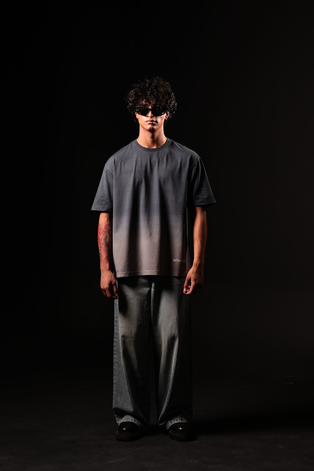 Person wearing a dark gray t-shirt and wide-leg pants with a black mask on a black background