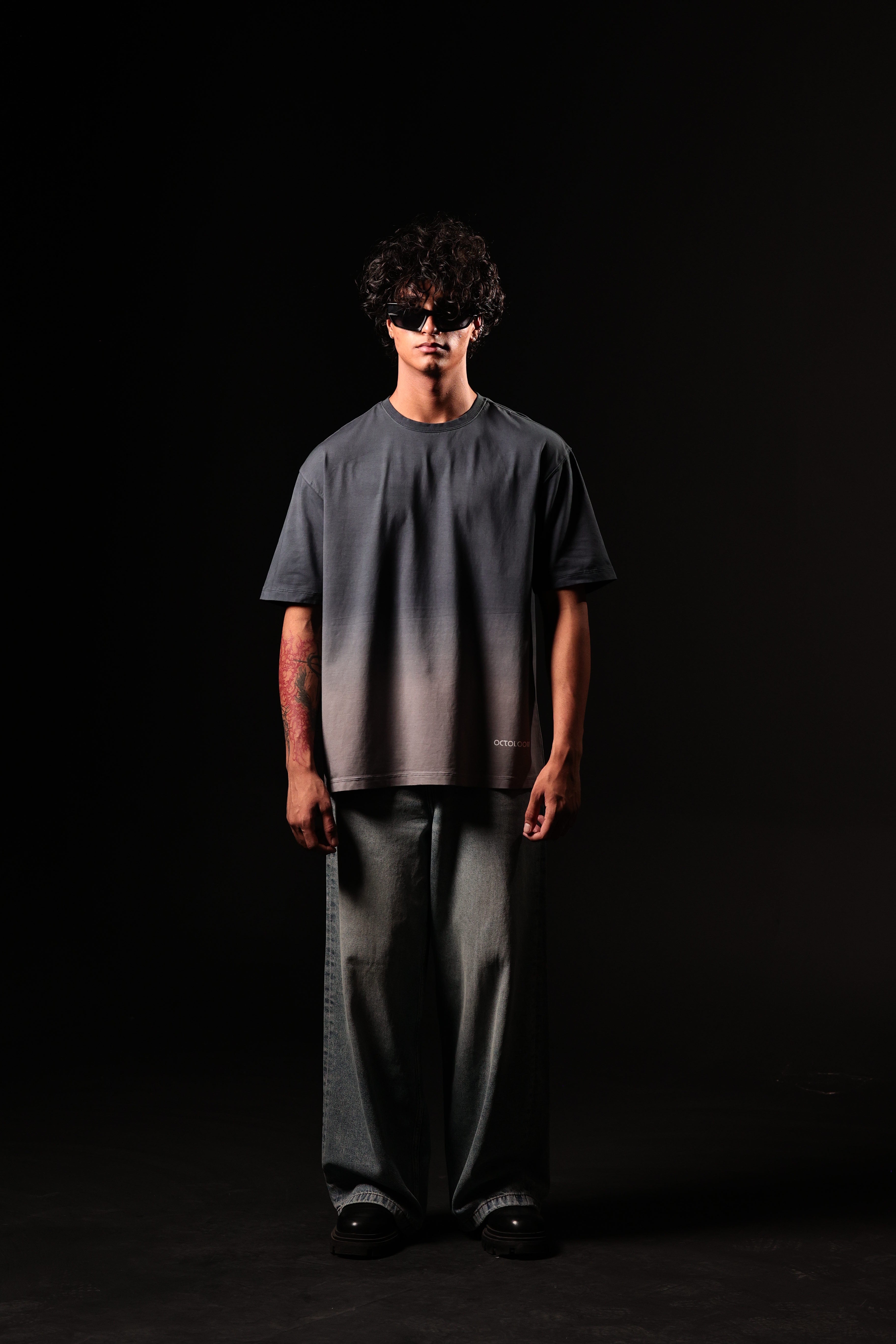 Person wearing a dark gray t-shirt and wide-leg pants with a black mask on a black background